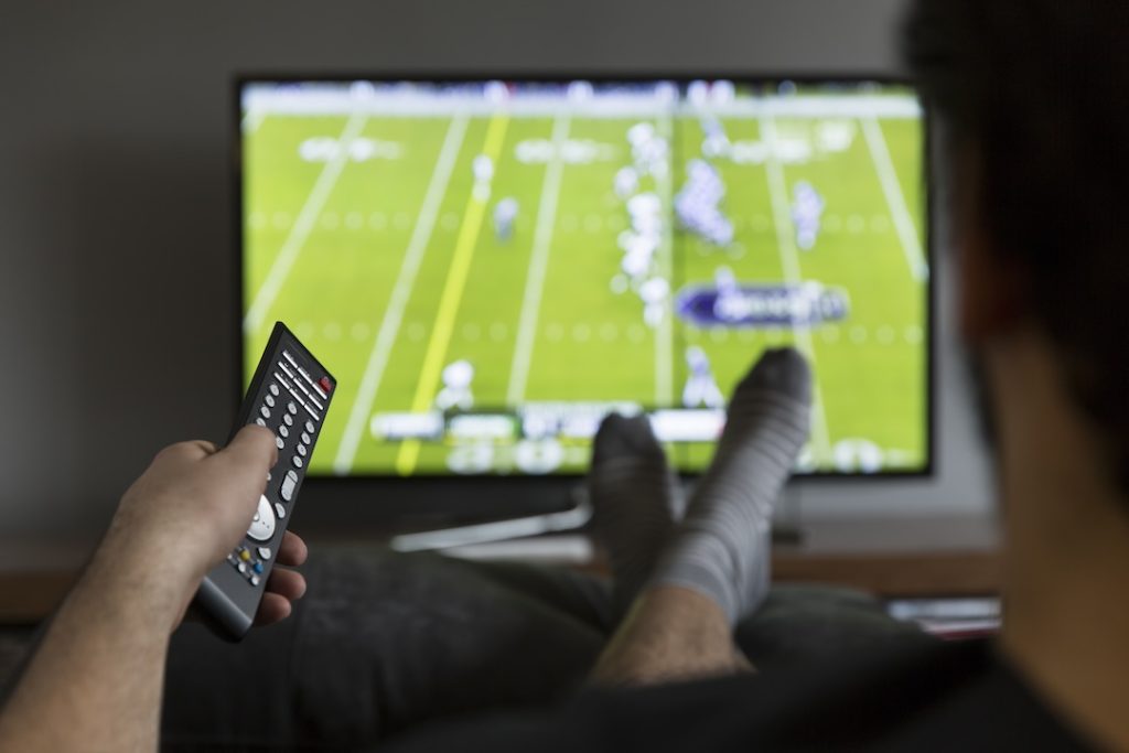 A man points a remote at the TV while watching a sports game with his feet up.