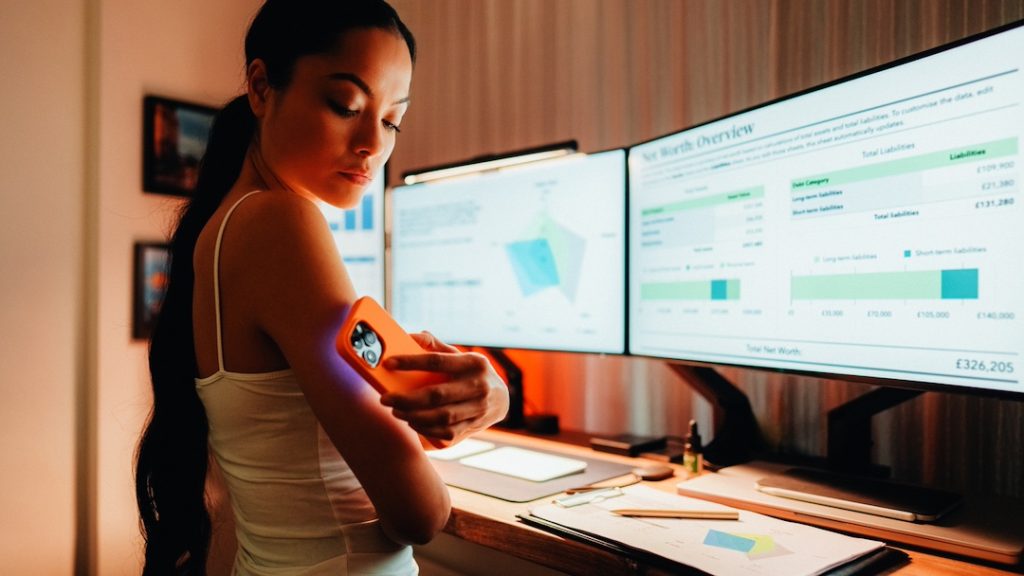 A young woman checks her glucose levels while at her desk working from home, showing how wearable tech devices range from smartwatches to glucose monitors for people with diabetes.